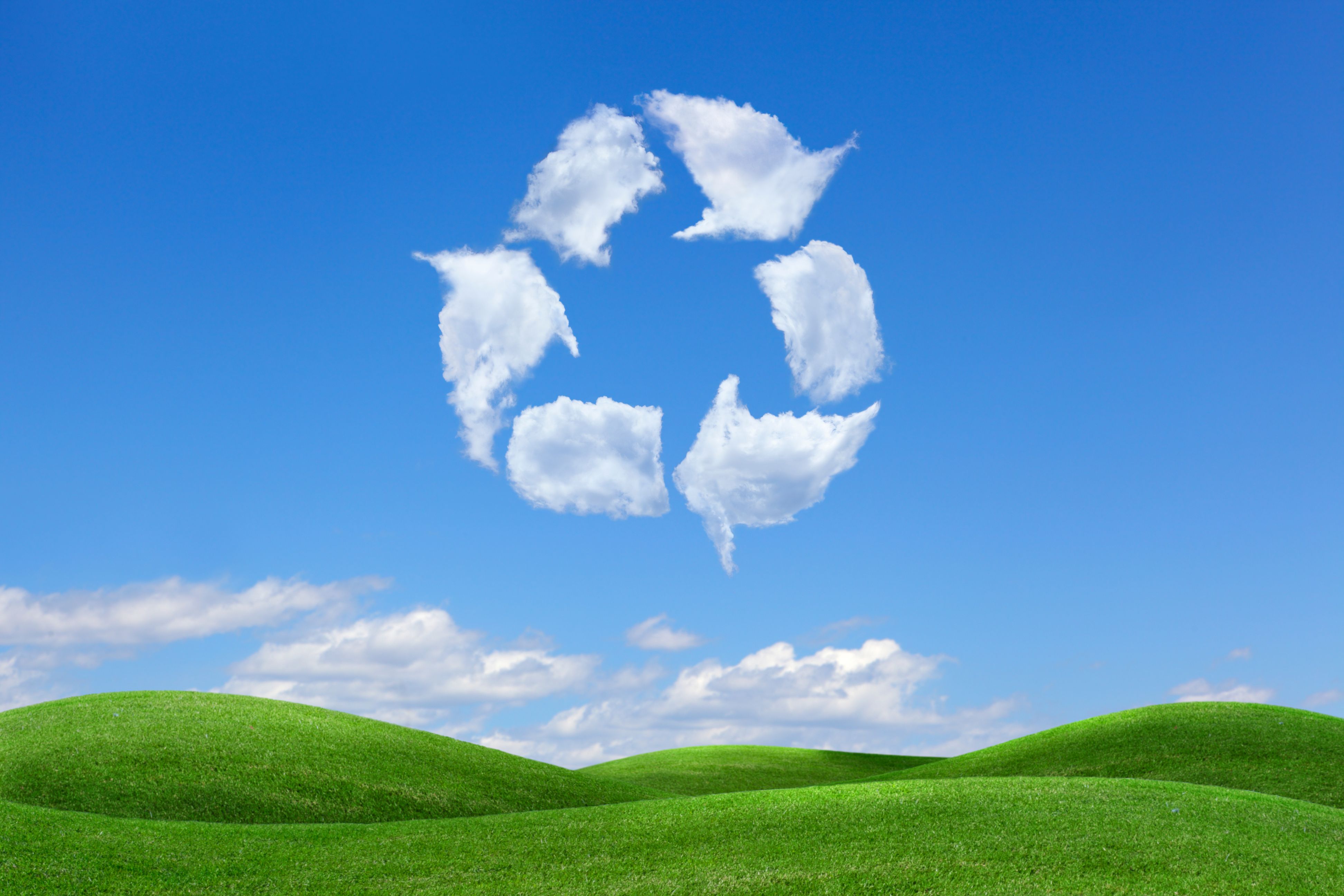 Recycling symbol looking like clouds, in the blue sky with green grass below.