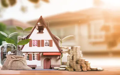 A tiny model house out front with a pile of money beside it, and a larger home blurred out in the background.