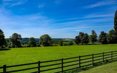 A farm fence, with a green field, rolling hills and trees behind.