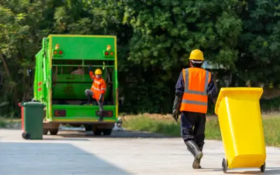 Garbage truck in the background with a worker beside it, and another worker in the foreground pulling a garbage can toward the truck.