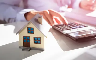 A model of a house, with a person using a calulator beside a pile of papers in the background.