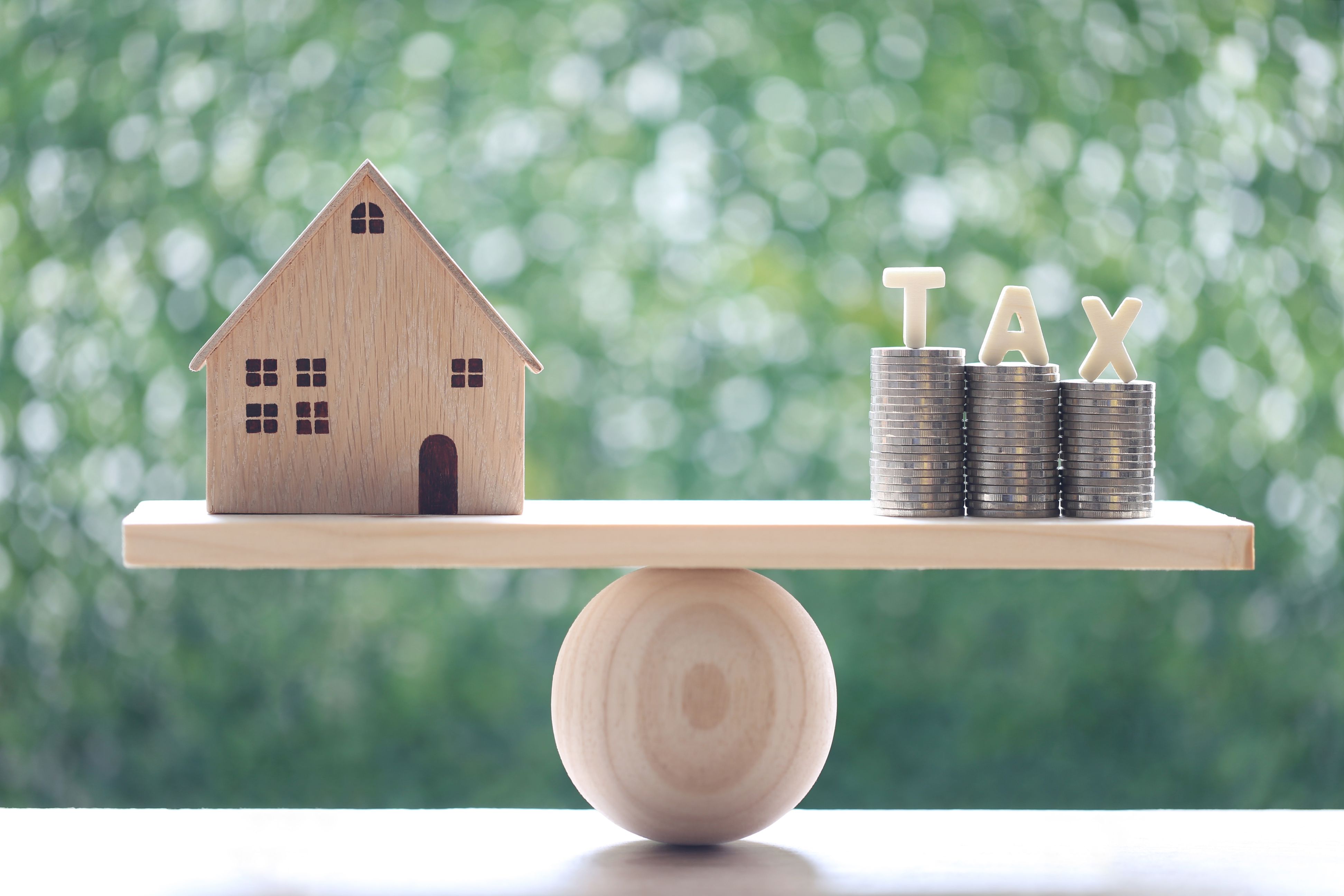 A wooden model house balancing on a wooden board laying on a wooden ball, with coins on the opposite side.