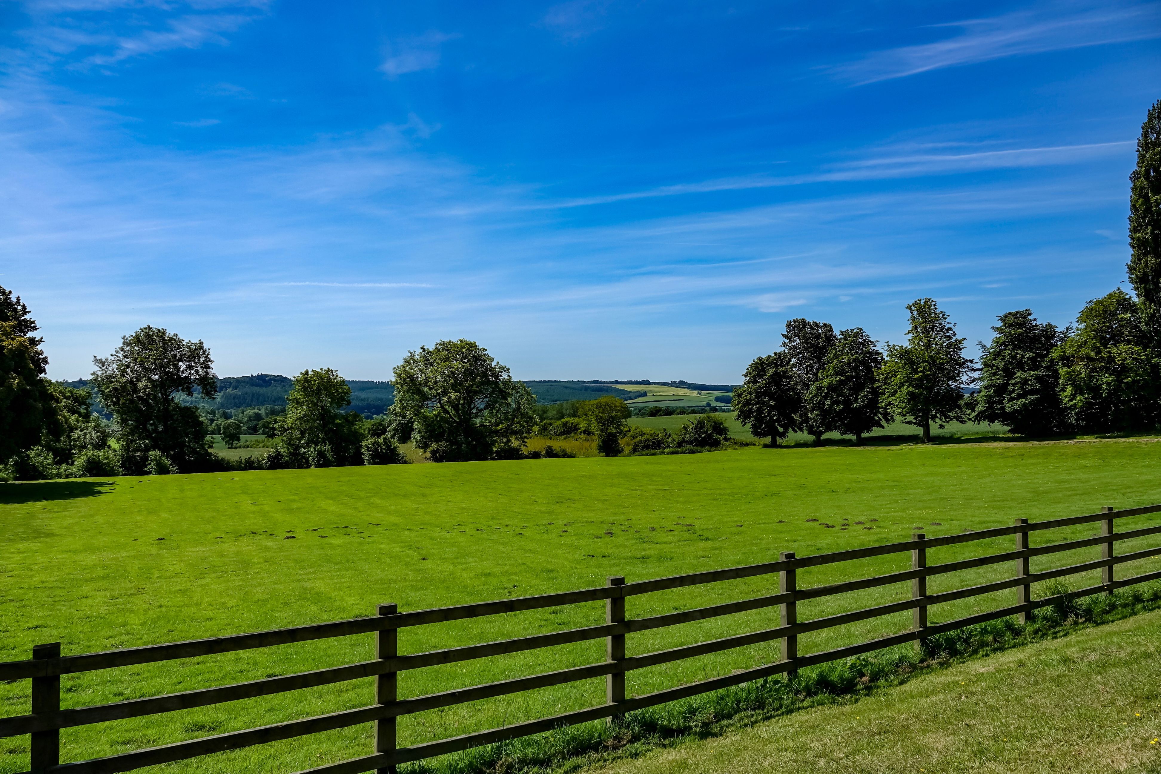 A farm fence, with a green field, rolling hills and trees behind.